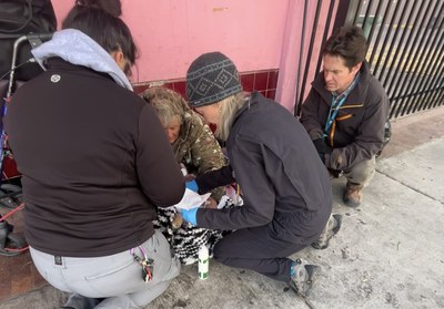 Members of the Street Medicine Team kneel on a sidewalk to assist an unsheltered individual. A healthcare worker wearing gloves examines the individual, while two ACS Street Outreach Responders provide support and documentation. The scene highlights compassionate care being delivered directly in the individual’s environment, demonstrating the collaborative effort between medical and social service teams.