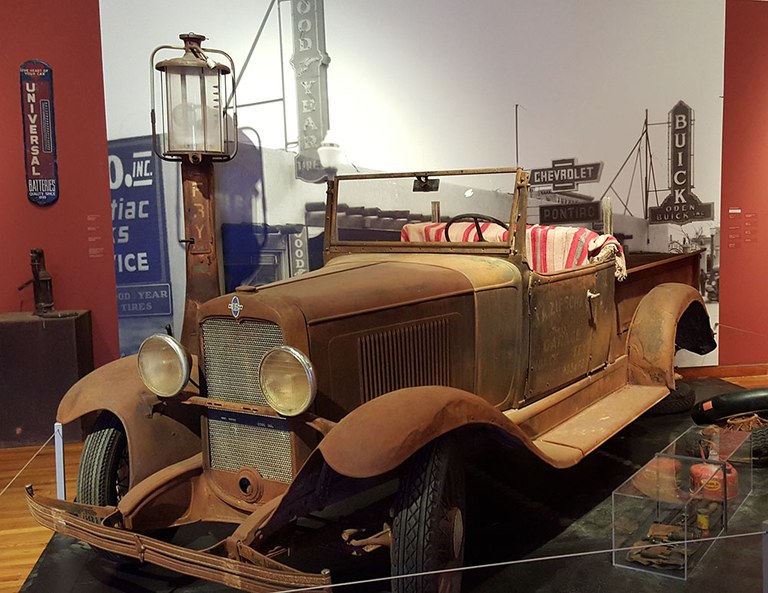 An antique car, brown with rust, inside an exhibition space.