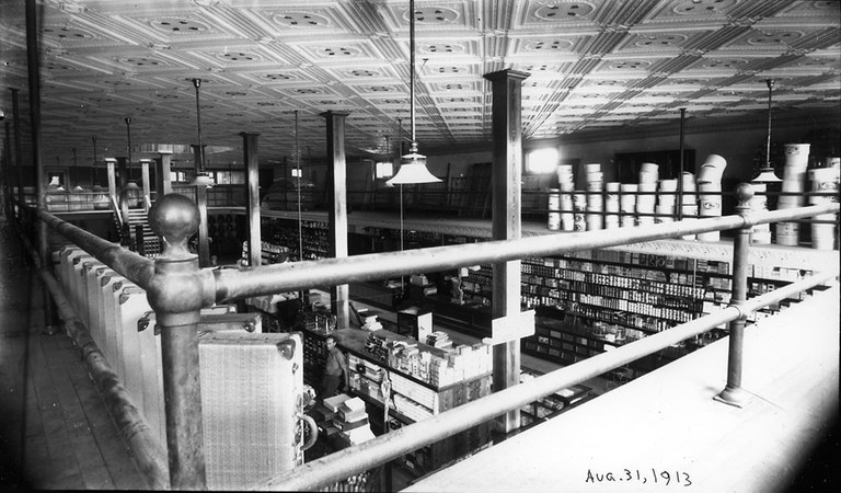 A vintage black and white photo of the inside of a store as seen from the upper level looking down. Some of the goods include suitcases and books.