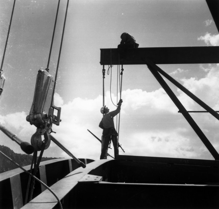A vintage black and white photo of a construction worker looking up and adjusting pullies on a crane.