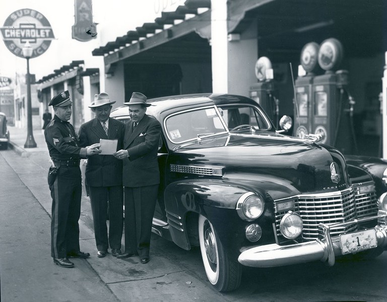 A vintage black and white photo of a group of men standing in front of a car at a gas station.