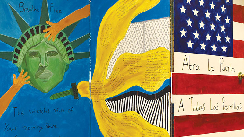 Photo of a brightly painted three-panel painted door with the head of Lady Liberty on a blue background and her torch oriented sideways in the middle panel and the American flag taking up the entire surface area of the third panel with the title of this work written on two of the white stripes on the flag: "Abra La Puerta" on one line which translates to "Open the Door" and below it "A Todas Las Familias" which translates to "For All the Families."