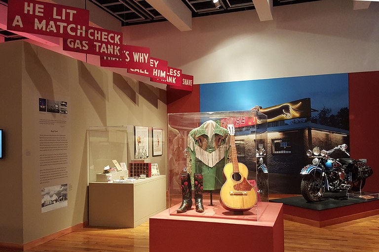 A portion of the Stories of the Road section of the Route 66 exhibition featuring a case with a guitar and boots and a motorcycle in the background.