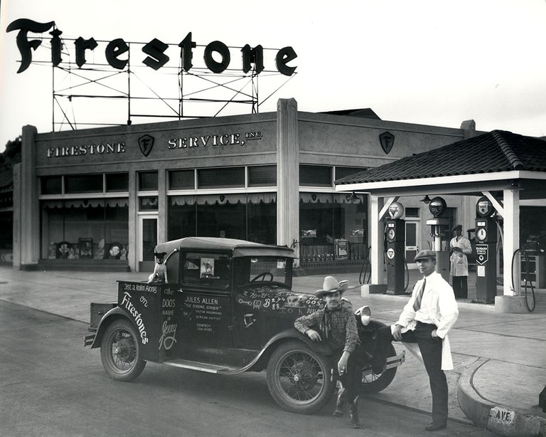A vintage black and white photo of a man standing next to a car on the road in front of a Firestone shop.
