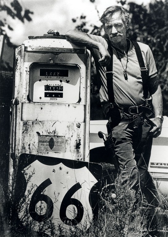 A black and white vintage photo of a man leaning on an old gas pump with a Route 66 sign leaning against it.