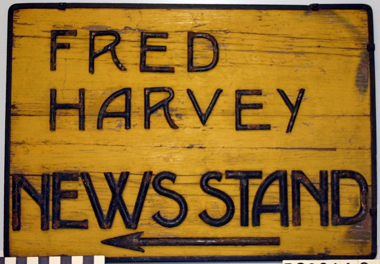 A wooden sign painted yellow with the text "Fred Harvey News Stand" in brown lettering.