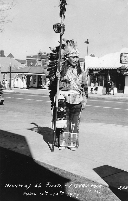 A black and white photo of a man wearing Native American attire standing on a sidewalk.
