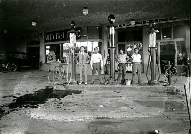 A black and white photo of a group of people posing in a line among the old fuel pumps at a gas station.