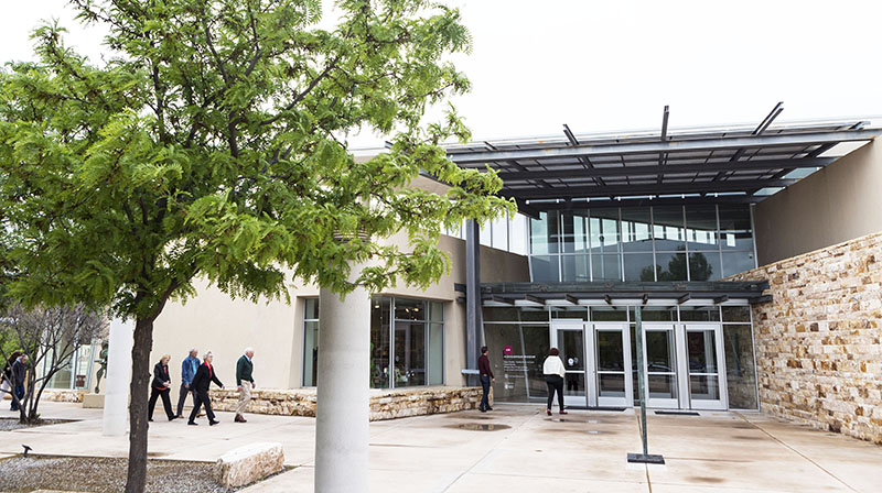 Photo of the front entrance of the museum with a tree in the left foreground and six people walking towards the front doors.