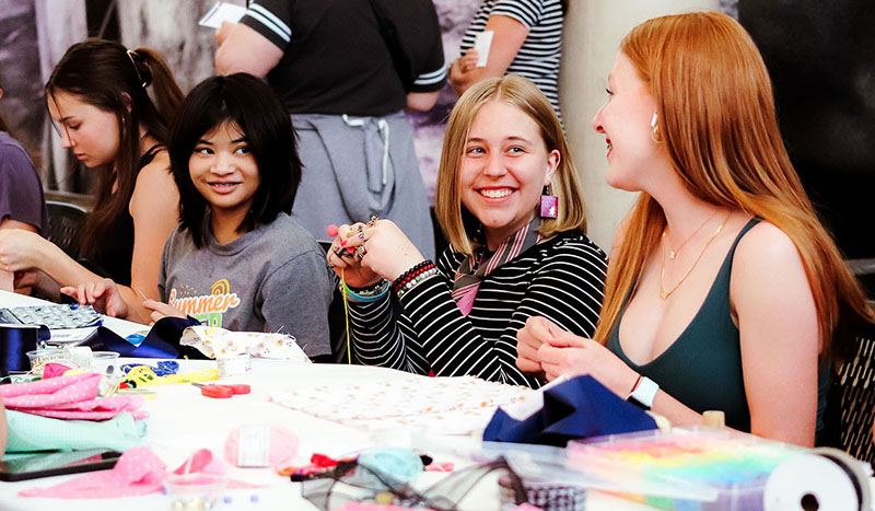 Photo of four girls seated at a rectangular table working on a textile art activity and looking at one another.