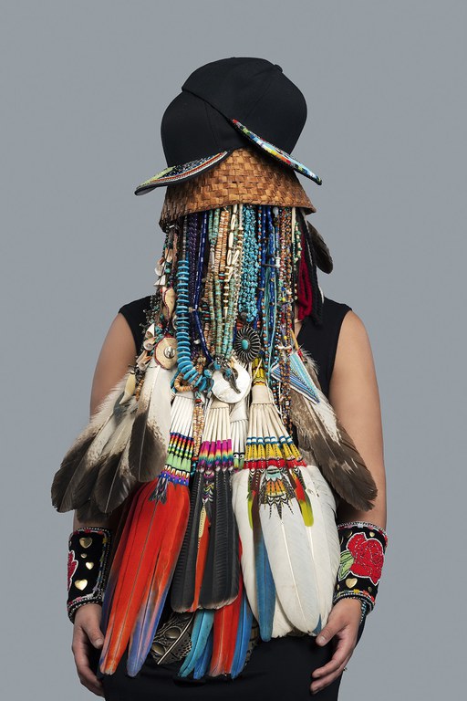 A woman poses with a costume made of multiple turquoise necklaces over her face and large bird feather over her torso. Headdress–Shadae, 2018