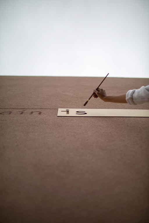 An intensely angled shot, taken from below, cropped tight over a hand holding a paintbrush up to a large letter stencil attached to an adobe wall.