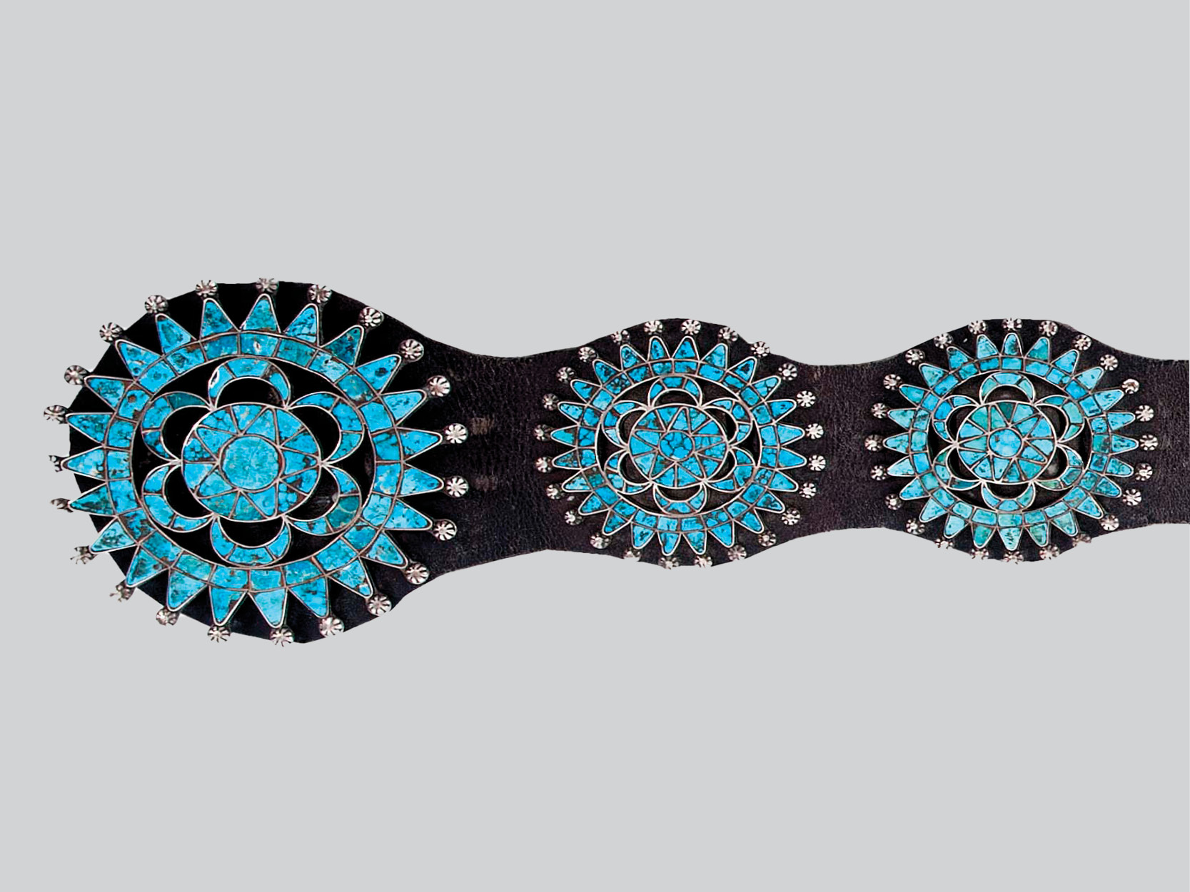 Detail of a concha belt with a dark leather strap embellished with three round designs with rays projecting from the center all inlaid with blue turquoise and round embossed silver studs at each point.