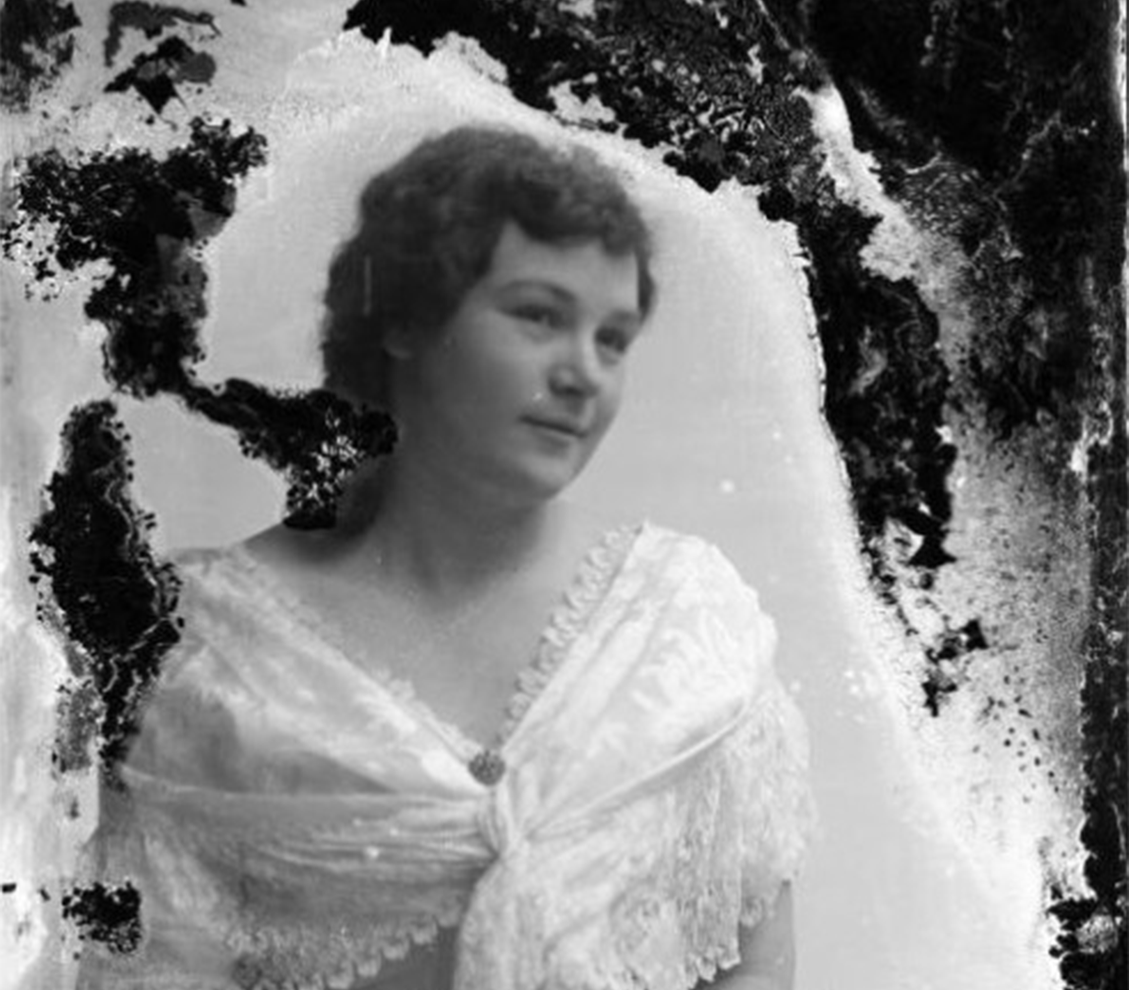 Portrait of a woman wearing a white top and a dark skirt. She has a lace shawl over her shoulders. Her hands are in her lap. She has dark hair pulled back from her face and is looking toward the right of the camera.