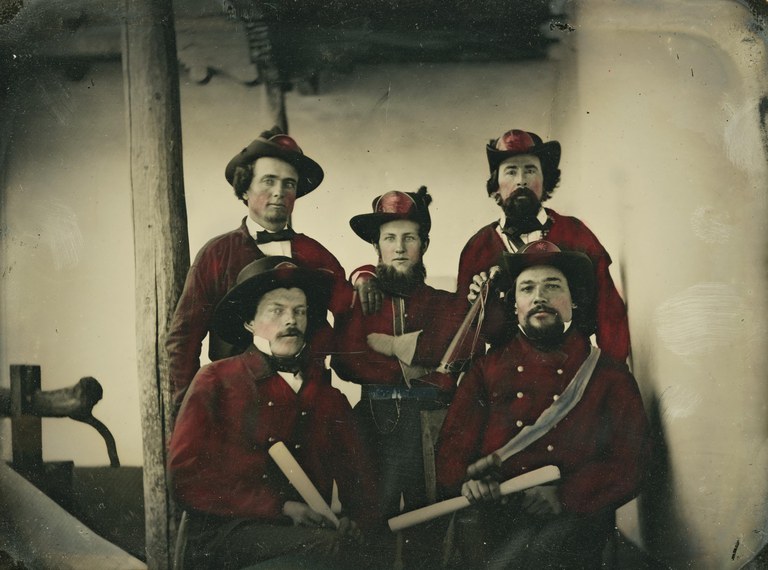 A black and white photo featuring five men of the Santa Fe Fire Department posing while wearing their uniforms. Their uniforms have been tinted red in a post-production artistic effect.