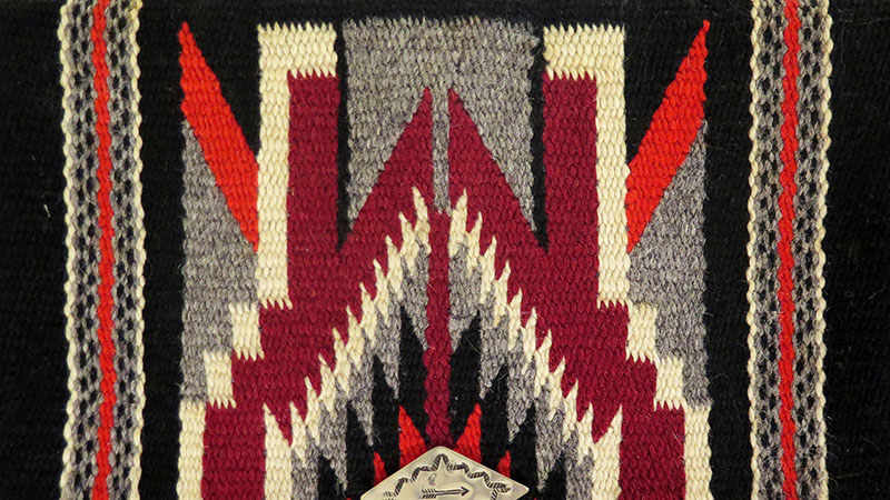 Museum collection photo of the front of a woven wallet with zigzag patterns and stripe patterns made from handloomed wool in burgundy, red, cream and black. Silver fastener at the bottom incised with a decorative pattern and an arrow in the center.