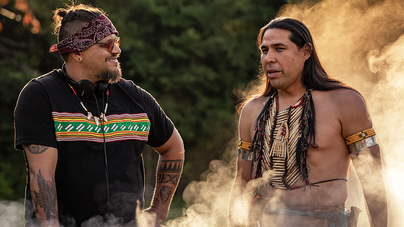 Photo of two Native American men talking outdoors shown from the waist up with some smoke effects rising from the bottom towards the upper right.