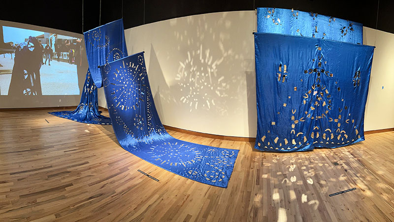 Photo of Tsedaye Makonnen’s installation of two royal blue textiles embellished with an ornate pattern of reflective glass and a mannequin wearing another version of the same in front of a video screen depicting the artist draped in the textile in an outdoor performance.