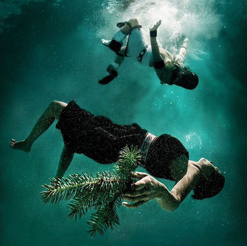 Underwater photograph of two clothed women floating on their backs in water with one more submerged holding an evergreen cutting in her left hand and the one above close to a white space that could be where she entered the water surface or it could be a source of light.