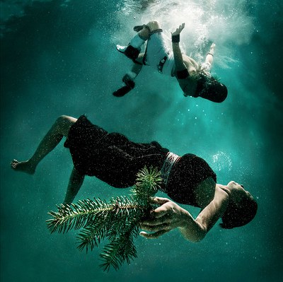 Underwater photograph of two clothed women floating on their backs in water with one more submerged holding an evergreen cutting in her left hand and the one above close to a white space that could be where she entered the water surface or it could be a source of light.