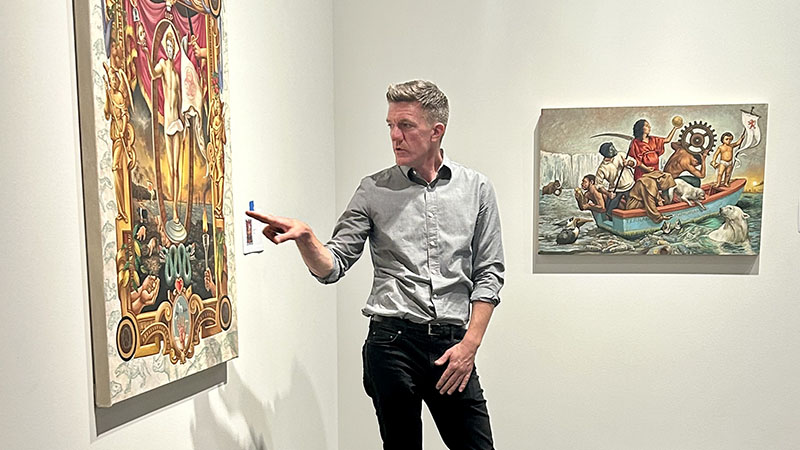 Photo of a man in a museum gallery pointing at a painting with another painting behind him.