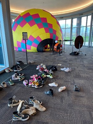 An RC hot air balloon, inflated and on its side, rests on the floor inside the Balloon Museum. In the foreground are dozens of shoes of children who are inside the RC balloon.