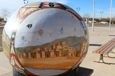 A large sculpture representing a Native American seed pot painted with a Pueblo scene with adobe buildings and hot air balloons flying overhead.