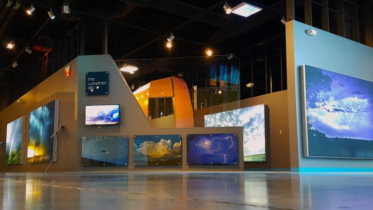 The Weather Lab as seen from outside the exhibition facing the wall with various large, colorful wall art pieces of different weather phenomenon.