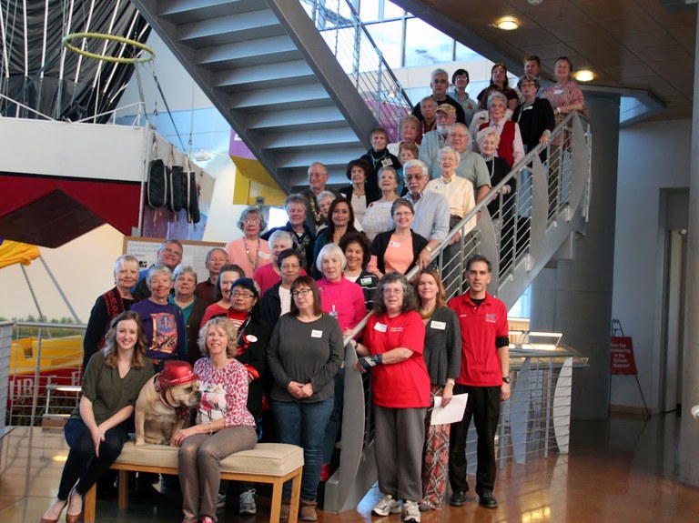 A large group of Balloon Museum volunteers gather on the stairs inside the Balloon Museum and pose, facing the audience.