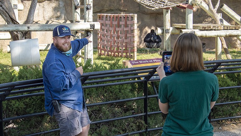 A keeper speaks and points at a gorilla while looking at another keeper who is recording him.