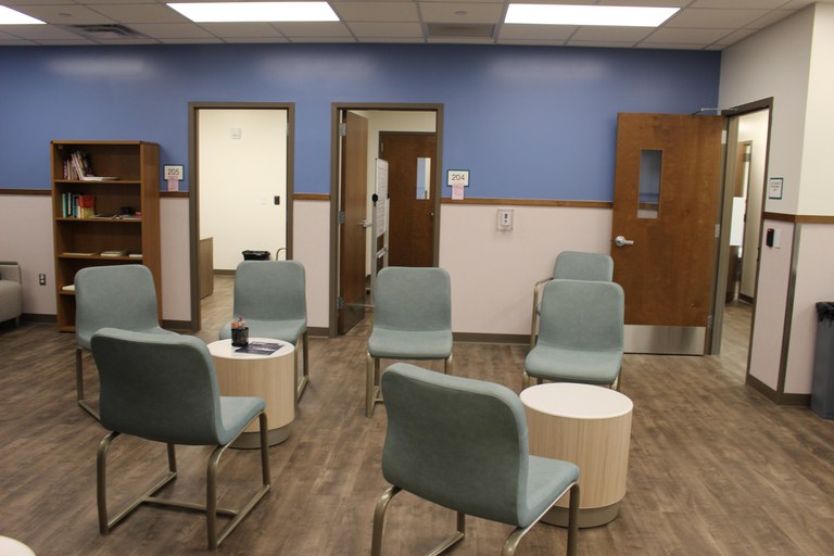 Two groups of four chairs are clustered around small tables. A bookcase has books. Open doors lead to offices. The color scheme is blue and soothing