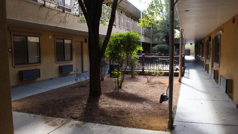 A walkway for motel housing that has sidewalk going around a square planting area with trees and dirt.