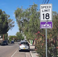 Shows the Silver Avenue bike boulevard with an 18 miles per hour speed limit sign with the purple bike boulevard sign underneath the speed limit sign. It also shows on-street parking that narrows the driving space.