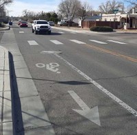 There is a bike lane adjacent to a sidewalk on one side and a vehicle driving lane on the other side. The bike lane has a bike lane symbol which shows a person on a bicycle. Where the bike lane crosses an intersection there is also a crosswalk. In the driving lane there are a few vehicles driving.