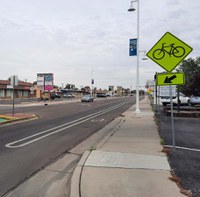 There is a bike lane with a striped buffer between the bike lane and the vehicle driving lane. There is also a bright yellow diamond-shaped sign with a bicycle symbol and an arrow pointing toward the bike lane. The sign is adjacent to the sidewalk.