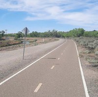 There is an asphalt paved multi-use trail that has a yellow dashed line in the middle of the paved area to indicate bi-directional travel for people walking and biking. In the distance coming toward the camera, there is a person bicycling.