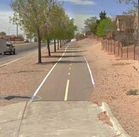 There is a two-lane roadway with a truck driving away from the camera. Next to the roadway is a landscaped area with many green trees and next to the landscaped area is an asphalt paved sidepath with a yellow dashed line delineating bi-directional traffic for people walking and biking. Next to the sidepath is a dirt area and then there are fences to people's backyards.