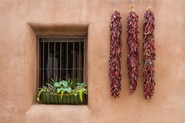 A stucco home close-up, featuring a window with metal bars and a potted plan on the sill. Three red chile ristras hang just to the right.