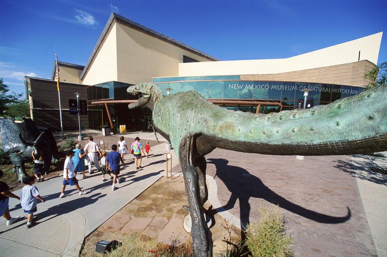 Front of the Museum of Natural History from the perspective of the tail area of a bronze dinosaur sculpture. Patrons walk toward the entrance in the background.