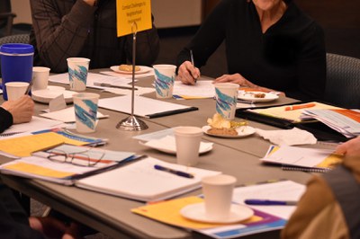 A close up of a gray conference table filled with notebooks, paper, and pens and several people writing.