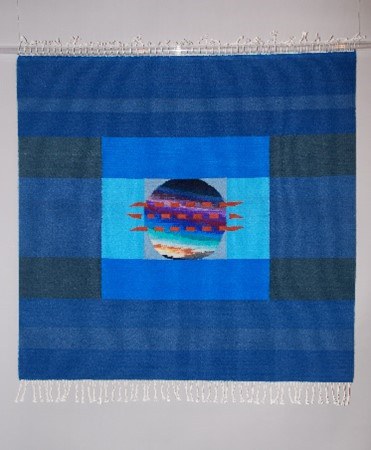 A geometric tapestry in shades of blue. A blue circle design is inside of a small blue square inside of a larger darker blue square with horizontal rows of dark and light color alternating from top to bottom.