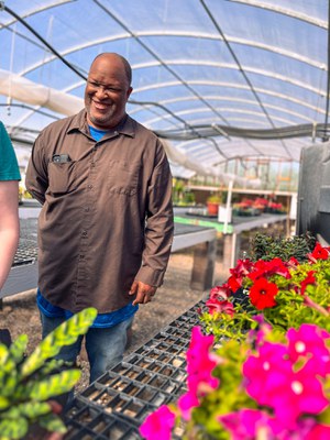 A volunteer standing next to a table of flowers smiling with his hand behind his back.