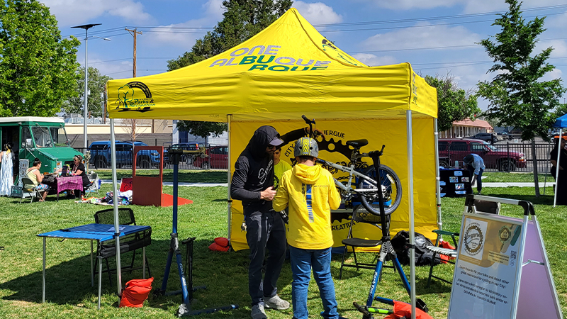 A staff member helping someone fix their bike at a park under a yellow tent with logos for Esperanza Bicycle Safety Education Center, OneAlbuquerque logo in green for the Parks and Recreation department.