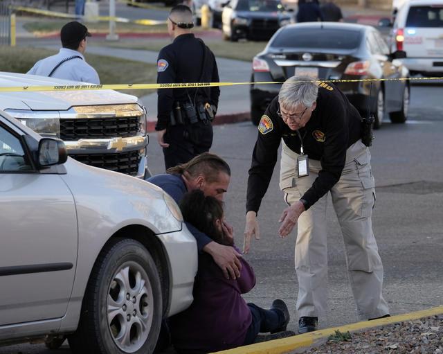 A Police chaplain bending over to talk to a woman and man hugging while sitting in front of a vehicle that has been taped off with crime scene tape.