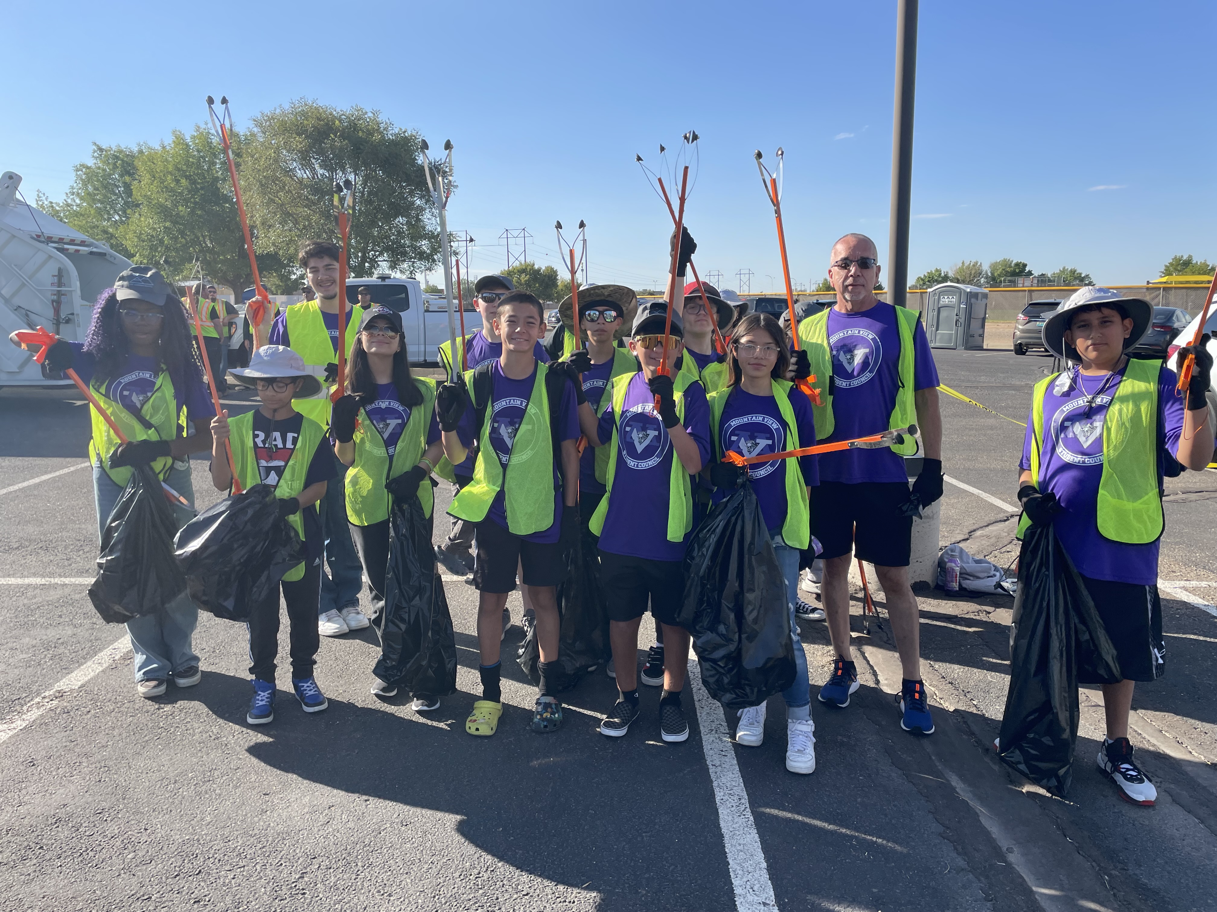 A group of adults stand in two rough rows and smile at the audience. Behind them is a Solid Waste trash truck with the One Albuquerque logo on the side. Bits of trees and a telephone pole can be seen in the background.