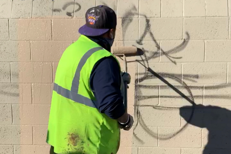 A city employee in a reflective vest painting over graffiti on a wall.
